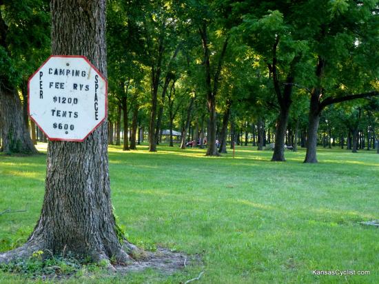 Neosho Falls Riverside Park - Fees - A sign showing the camping fees at Riverside Park. A family has set up their tent among the trees in the background.